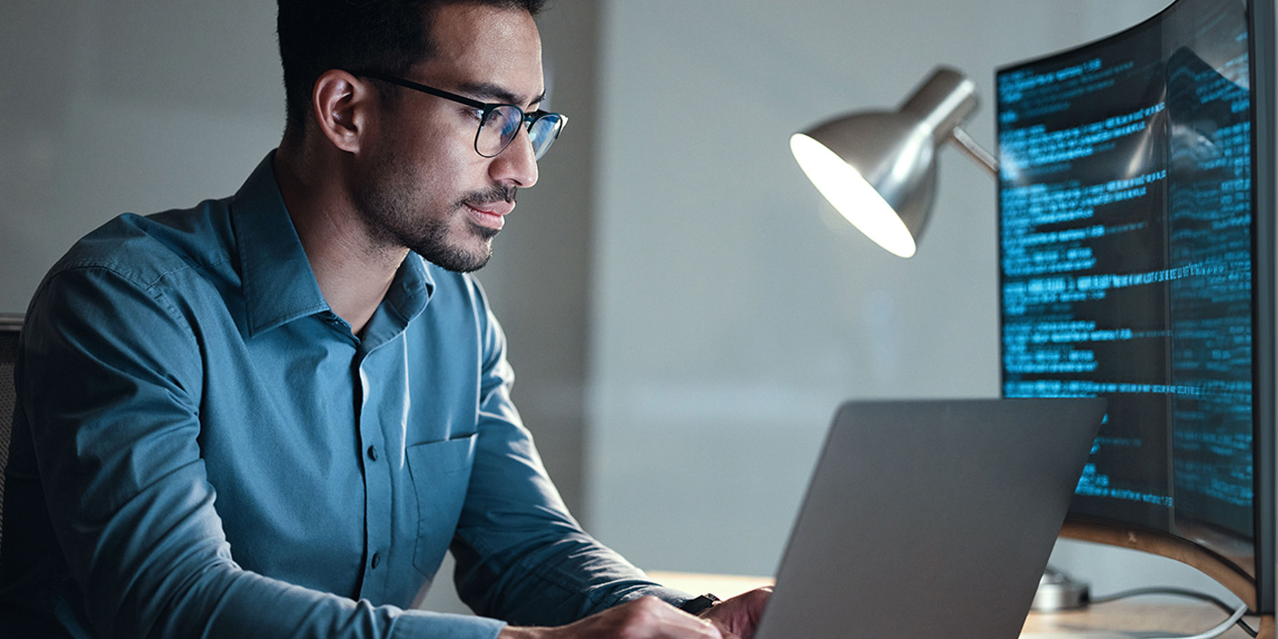 Stock image of a young man using a laptop while a screen in the back shows code