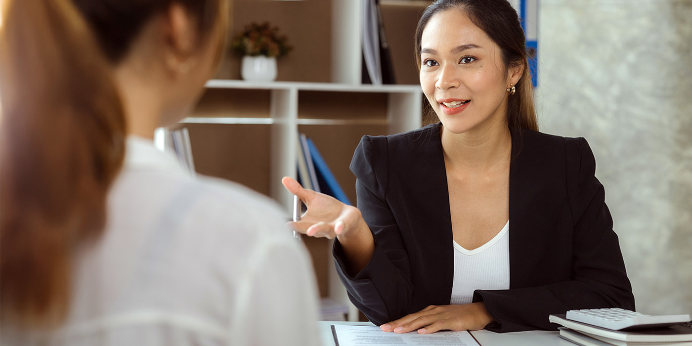 A stock photo of a business professional woman talking to another woman while both are sitting at a desk